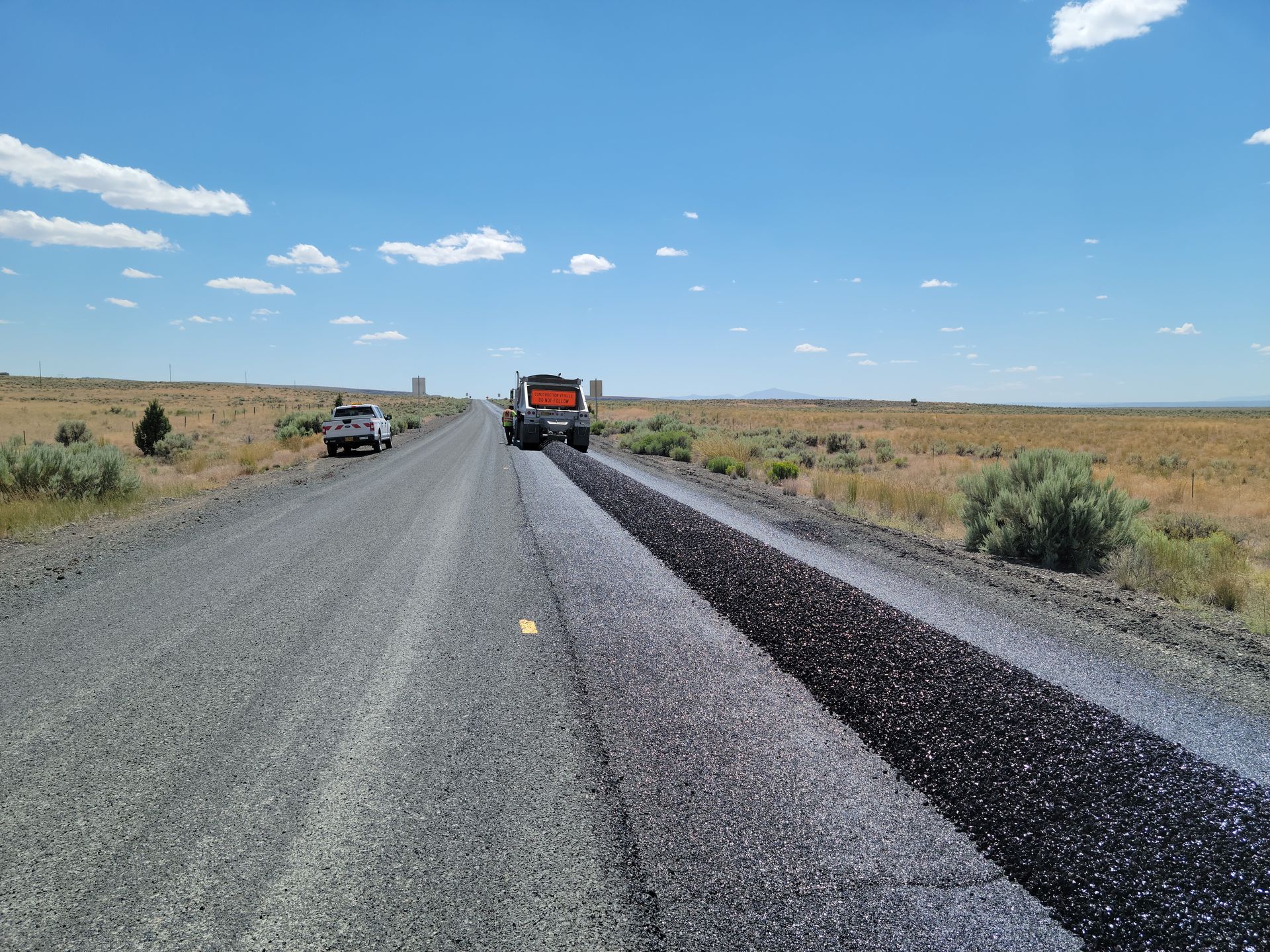 Construction equipment resurfacing a rural highway under a clear blue sky.