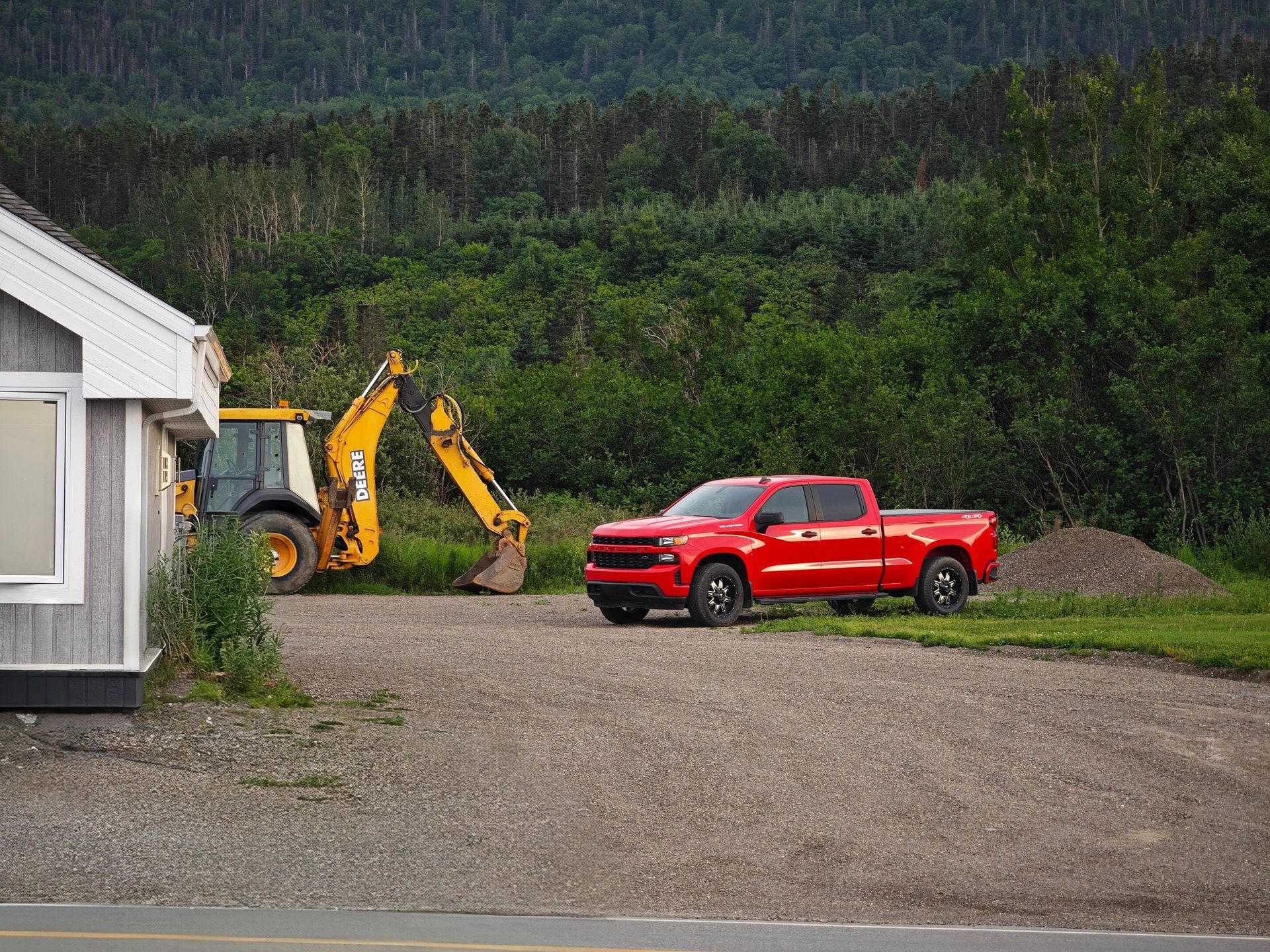 A red pickup truck and yellow backhoe parked in a rural driveway with lush forest backdrop.